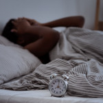 Sleepless, woman suffering from insomnia, sleep apnea or stress, headache or migraine. Tired and exhausted african american lady awake in middle of night, near alarm clock with time in dark, panorama