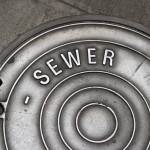 A sewer manhole cover in the sidewalk that is well worn and shiny