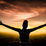 A young African American Woman raises her arms facing the sunset down over the valley and the ocean.