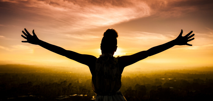 A young African American Woman raises her arms facing the sunset down over the valley and the ocean.