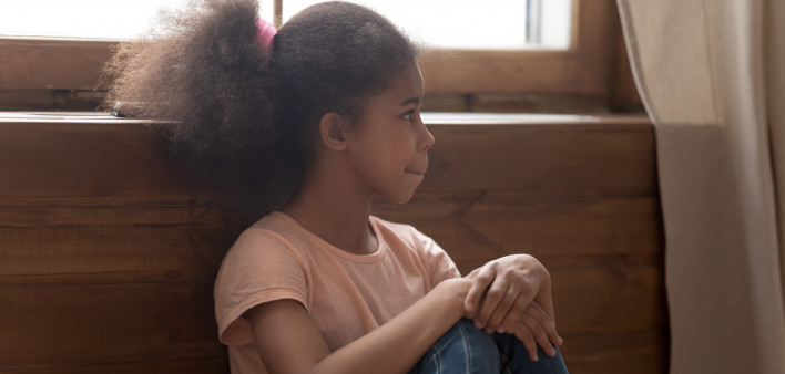 Pensive african preschool or school age girl sitting on floor alone, leaned on wooden wall puts hands on laps looking away