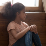 Pensive african preschool or school age girl sitting on floor alone, leaned on wooden wall puts hands on laps looking away