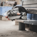 Poor homeless man or refugee sleeping on the wooden bench on the urban street in the city,