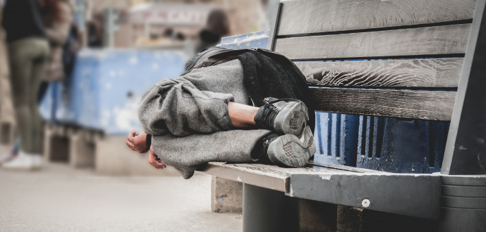 Poor homeless man or refugee sleeping on the wooden bench on the urban street in the city,