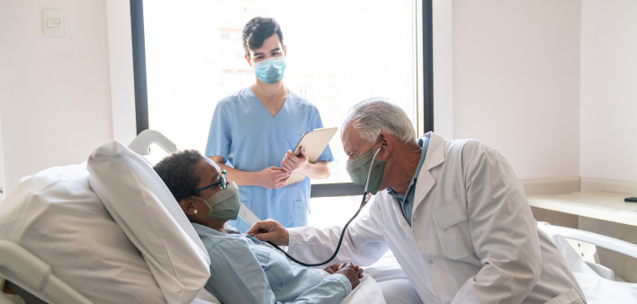 Senior male doctor checking black hospitalized female patient listening to her heart and nurse standing next to them all wearing protective facemasks