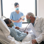 Senior male doctor checking black hospitalized female patient listening to her heart and nurse standing next to them all wearing protective facemasks