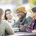 Five young adults are sitting at a picnic table outside on a fall day. They are smiling and chatting