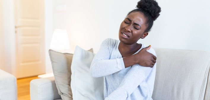 African woman sitting on couch take break touches her shoulder suffers from painful feelings ache caused by poor wrong posture, sedentary work, sitting at laptop for long period