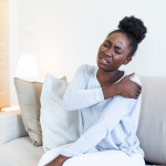 African woman sitting on couch take break touches her shoulder suffers from painful feelings ache caused by poor wrong posture, sedentary work, sitting at laptop for long period