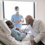 Senior male doctor checking black hospitalized female patient listening to her heart and nurse standing next to them all wearing protective facemasks