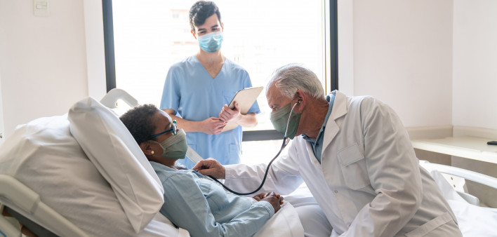 Senior male doctor checking black hospitalized female patient listening to her heart and nurse standing next to them all wearing protective facemasks