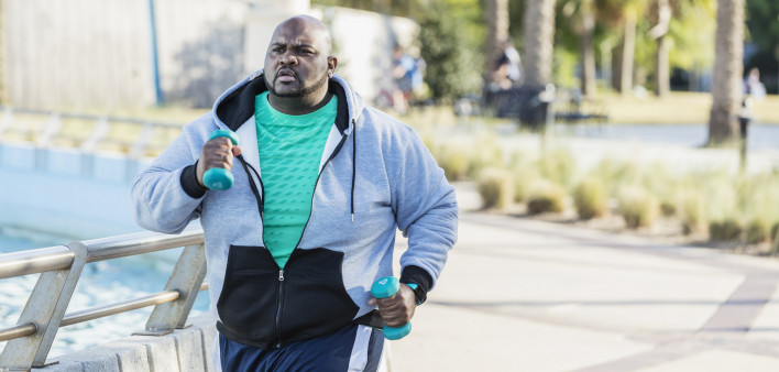 A mature African-American man in his 40s with a large build, exercising outdoors in a city park. He is wearing a fitness tracker, jogging or power walking with hand weights.