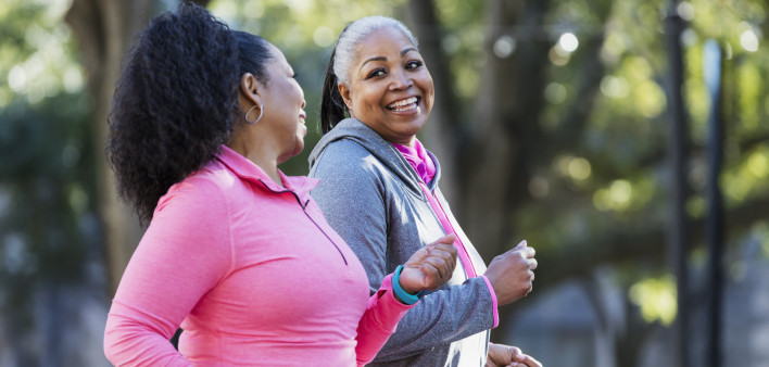 Two African-American women exercising together in the city, jogging or power walking, laughing and conversing. Buildings and trees are out of focus in the background. The one in pink is in her 60s...