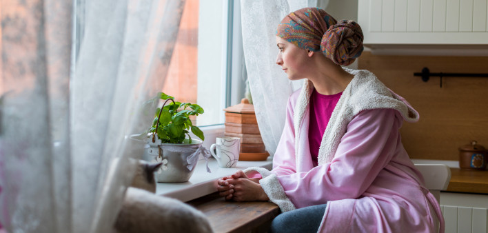 Young adult female cancer patient wearing headscarf and bathrobe sitting in the kitchen with her pet cat, looking out window.