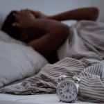 Sleepless, woman suffering from insomnia, sleep apnea or stress, headache or migraine. Tired and exhausted african american lady awake in middle of night, near alarm clock with time in dark, panorama