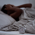 Sleepless, woman suffering from insomnia, sleep apnea or stress, headache or migraine. Tired and exhausted african american lady awake in middle of night, near alarm clock with time in dark, panorama