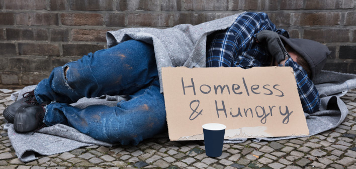 Male Beggar Lying On Street With Homeless And Hungry Text On Cardboard