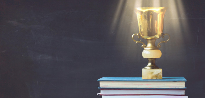Golden trophy on pile of books, against blackboard, with sun rays over trophy; learning/achievement