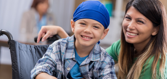 Mid adult mom and her elementary age son wait in waiting room in doctor's office or hospital. The boy is wearing a blue cap and is sitting in a wheelchair.