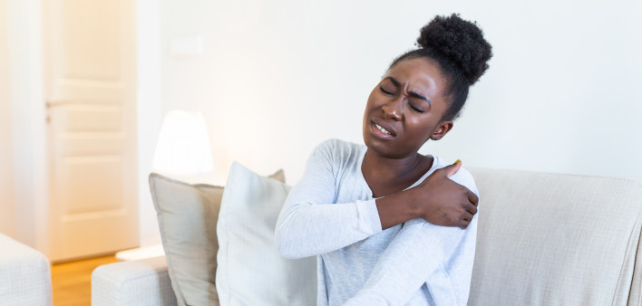 African woman sitting on couch take break touches her shoulder suffers from painful feelings ache caused by poor wrong posture, sedentary work, sitting at laptop for long period