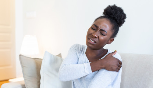 African woman sitting on couch take break touches her shoulder suffers from painful feelings ache caused by poor wrong posture, sedentary work, sitting at laptop for long period