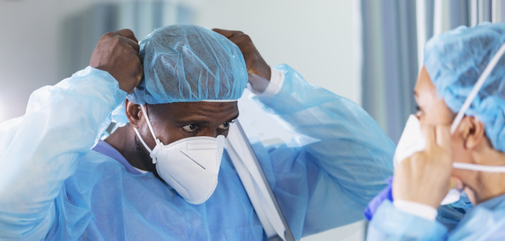 Two multiracial healthcare workers working in a hospital ward, putting on full PPE. They are wearing surgical caps and gowns and adjusting their N95 face masks.