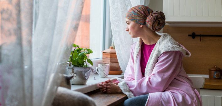 Young adult female cancer patient wearing headscarf and bathrobe sitting in the kitchen with her pet cat, looking out window.