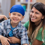 Mid adult mom and her elementary age son wait in waiting room in doctor's office or hospital. The boy is wearing a blue cap and is sitting in a wheelchair.