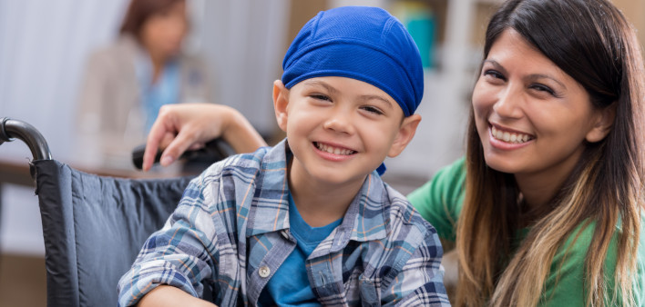 Mid adult mom and her elementary age son wait in waiting room in doctor's office or hospital. The boy is wearing a blue cap and is sitting in a wheelchair.