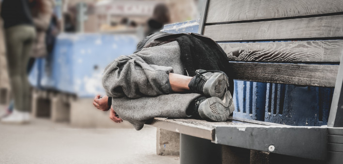 Poor homeless man or refugee sleeping on the wooden bench on the urban street in the city,