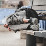 Poor homeless man or refugee sleeping on the wooden bench on the urban street in the city,