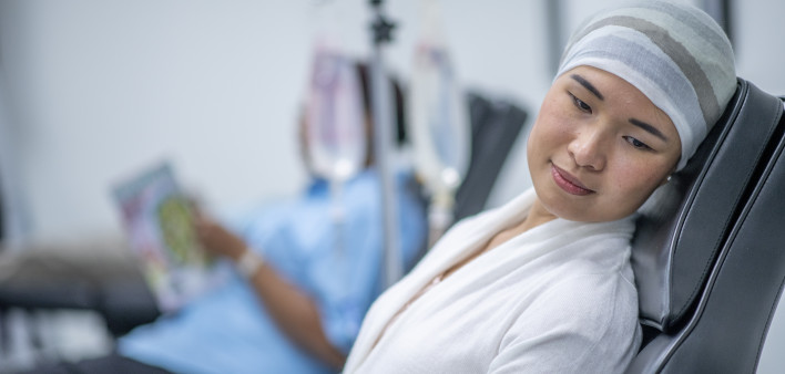 A woman wearing a head scarf lies on a hospital bed and looks to the side in contemplation. She is wearing a head scarf and a hospital gown and there is a IV drip next to her.