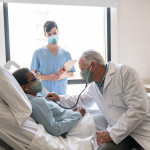 Senior male doctor checking black hospitalized female patient listening to her heart and nurse standing next to them all wearing protective facemasks