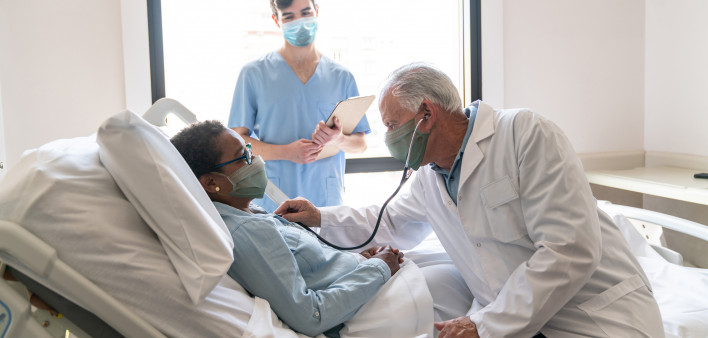 Senior male doctor checking black hospitalized female patient listening to her heart and nurse standing next to them all wearing protective facemasks