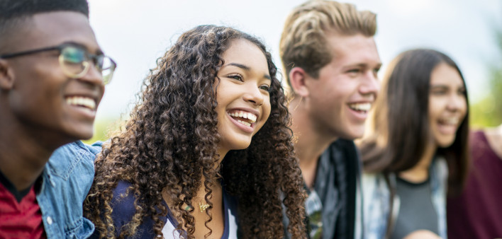 A girl of African American descent smiles with laughter while sitting in a row of teens with the arms around each other's shoulders. They are laughing too.