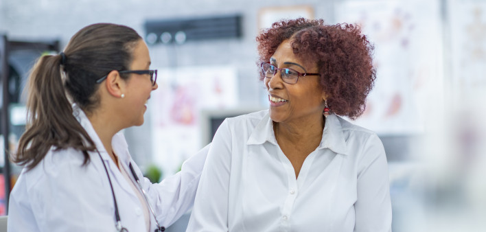 An African American woman smiles lightly as she discusses with the doctor sitting beside her. They are in a medical office.