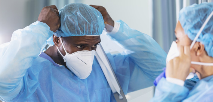 Two multiracial healthcare workers working in a hospital ward, putting on full PPE. They are wearing surgical caps and gowns and adjusting their N95 face masks.