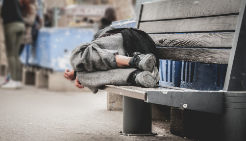 Poor homeless man or refugee sleeping on the wooden bench on the urban street in the city,