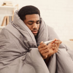 Ill african-american man covered with blanket holding thermometer, having fever