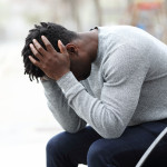 Side view portrait of a sad depressed black man sitting on a bench in a park