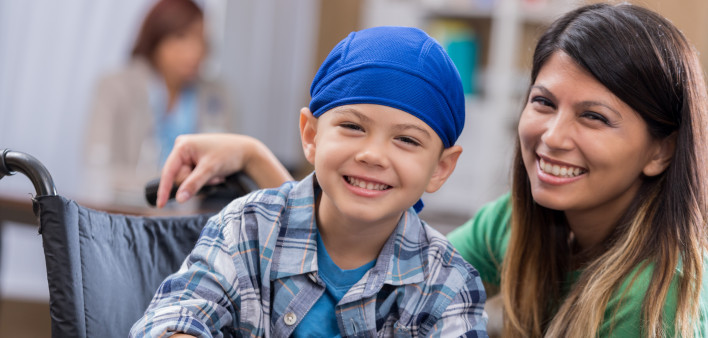 Mid adult mom and her elementary age son wait in waiting room in doctor's office or hospital. The boy is wearing a blue cap and is sitting in a wheelchair.
