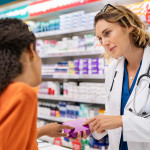 Pharmacist giving medicine box to customer in pharmacy. Doctor showing and explaining medicine dose to customer. Mature pharmacist giving advice on medicaments, while serving young african woman.