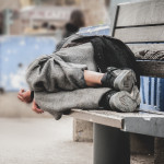 Poor homeless man or refugee sleeping on the wooden bench on the urban street in the city,