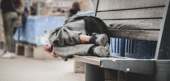 Poor homeless man or refugee sleeping on the wooden bench on the urban street in the city,