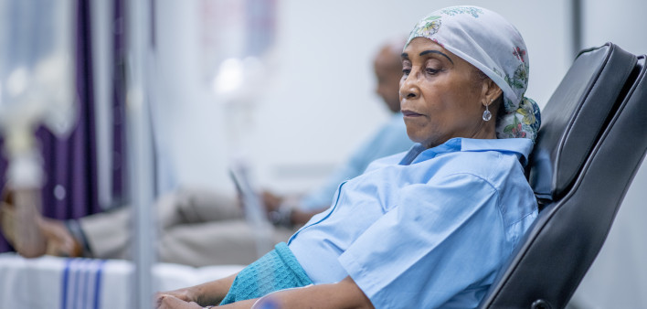 A woman wearing a head scarf lies on a hospital bed and looks to the side in contemplation.