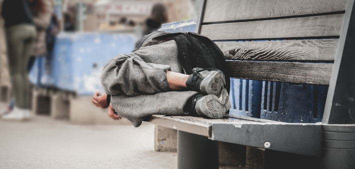 Poor homeless man or refugee sleeping on the wooden bench on the urban street in the city,