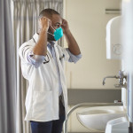 Side view of teenage African male medical student wearing short sleeved lab coat and looking in mirror as he puts on N95 face mask.