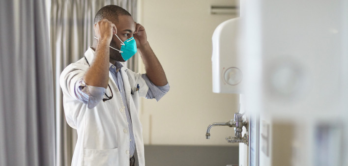 Side view of teenage African male medical student wearing short sleeved lab coat and looking in mirror as he puts on N95 face mask.
