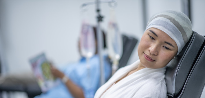 A woman wearing a head scarf lies on a hospital bed and looks to the side in contemplation. She is wearing a head scarf and a hospital gown and there is a IV drip next to her.