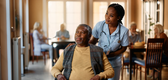 African American senior in wheelchair and young nurse looking through the window at residential care home.